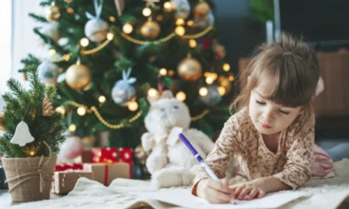 Ünnepi hangulatban kislány rajzol egy karácsonyfa előtt.